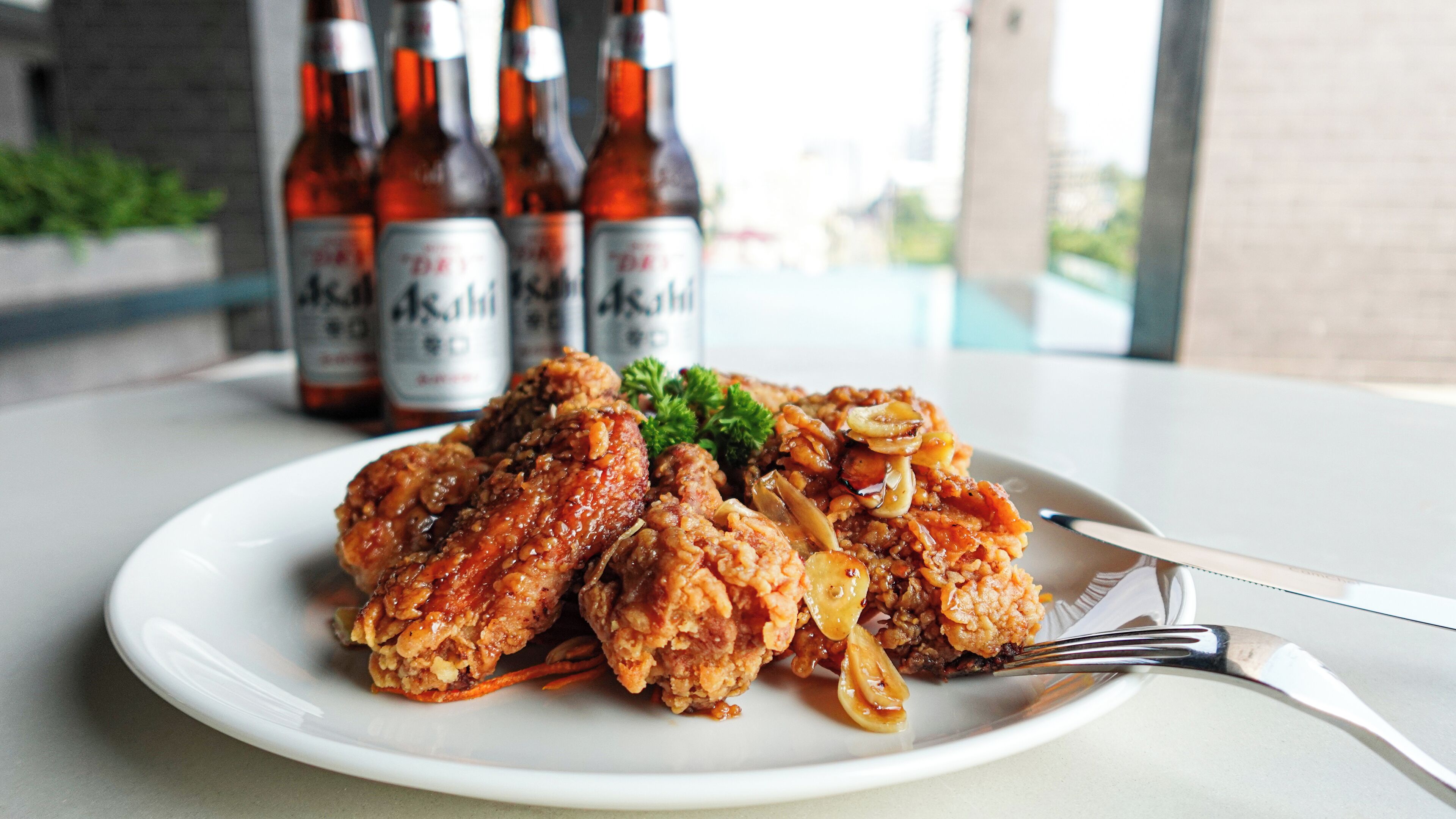 A plate of fried chicken with mushrooms and a garnish, accompanied by a fork and knife on a white plate. In the background, there are bottles of beer.