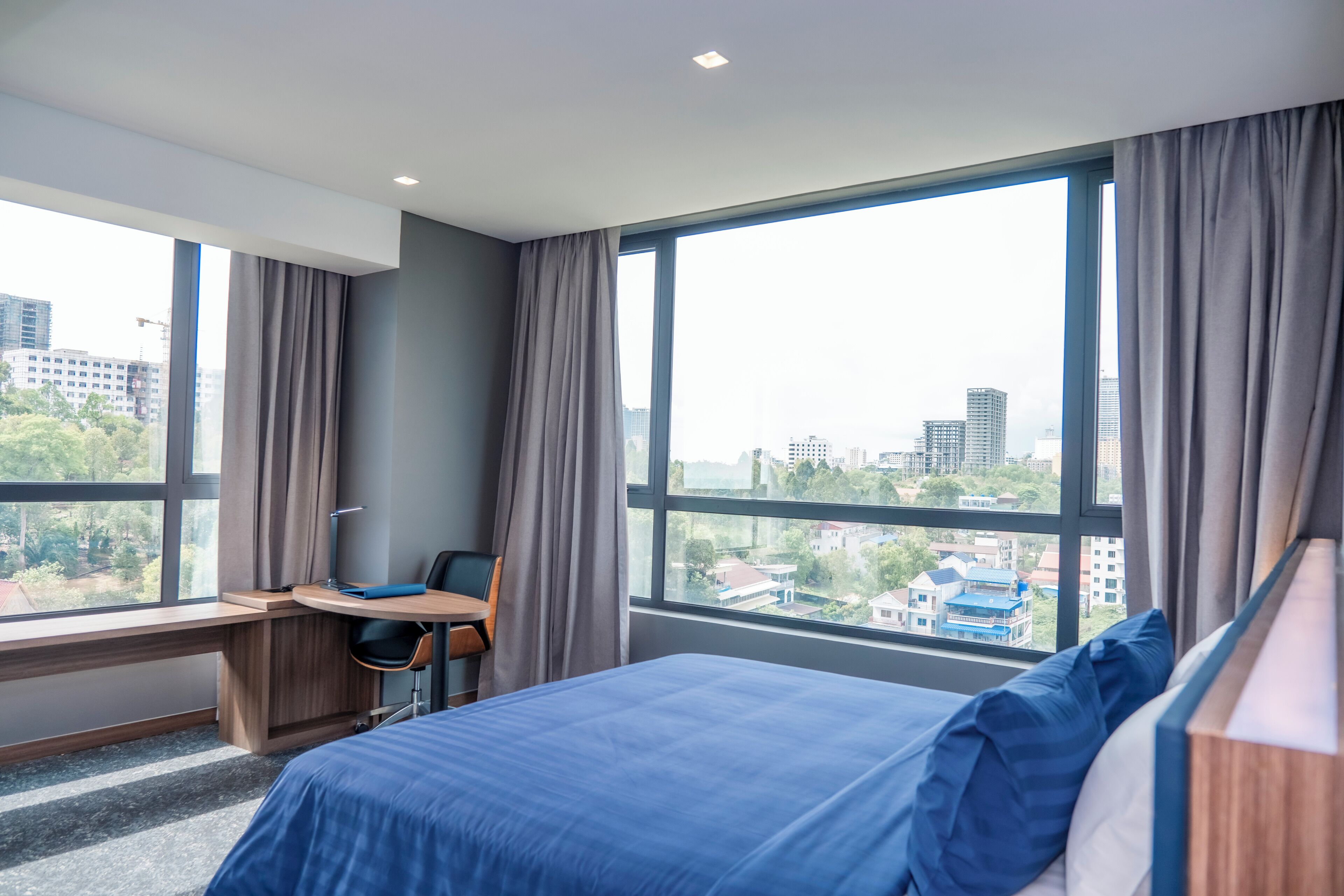 Une chambre d’hôtel moderne, dotée d’un grand lit, d’un bureau et offrant une vue sur la ville grâce à de grandes fenêtres.