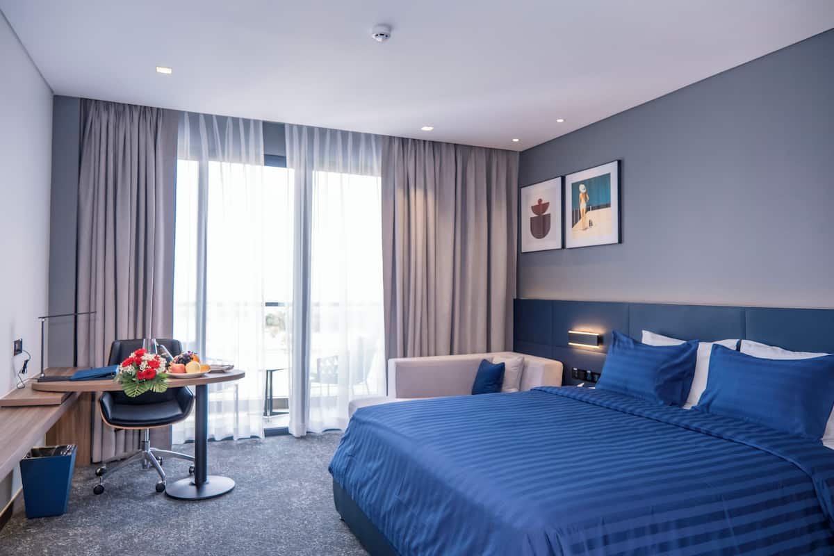 Ein Hotelzimmer mit einem großen Bett, einem Schreibtisch mit Stuhl, einem kleinen Tisch mit Blumenarrangement und Blick ins Freie durch ein großes Fenster.