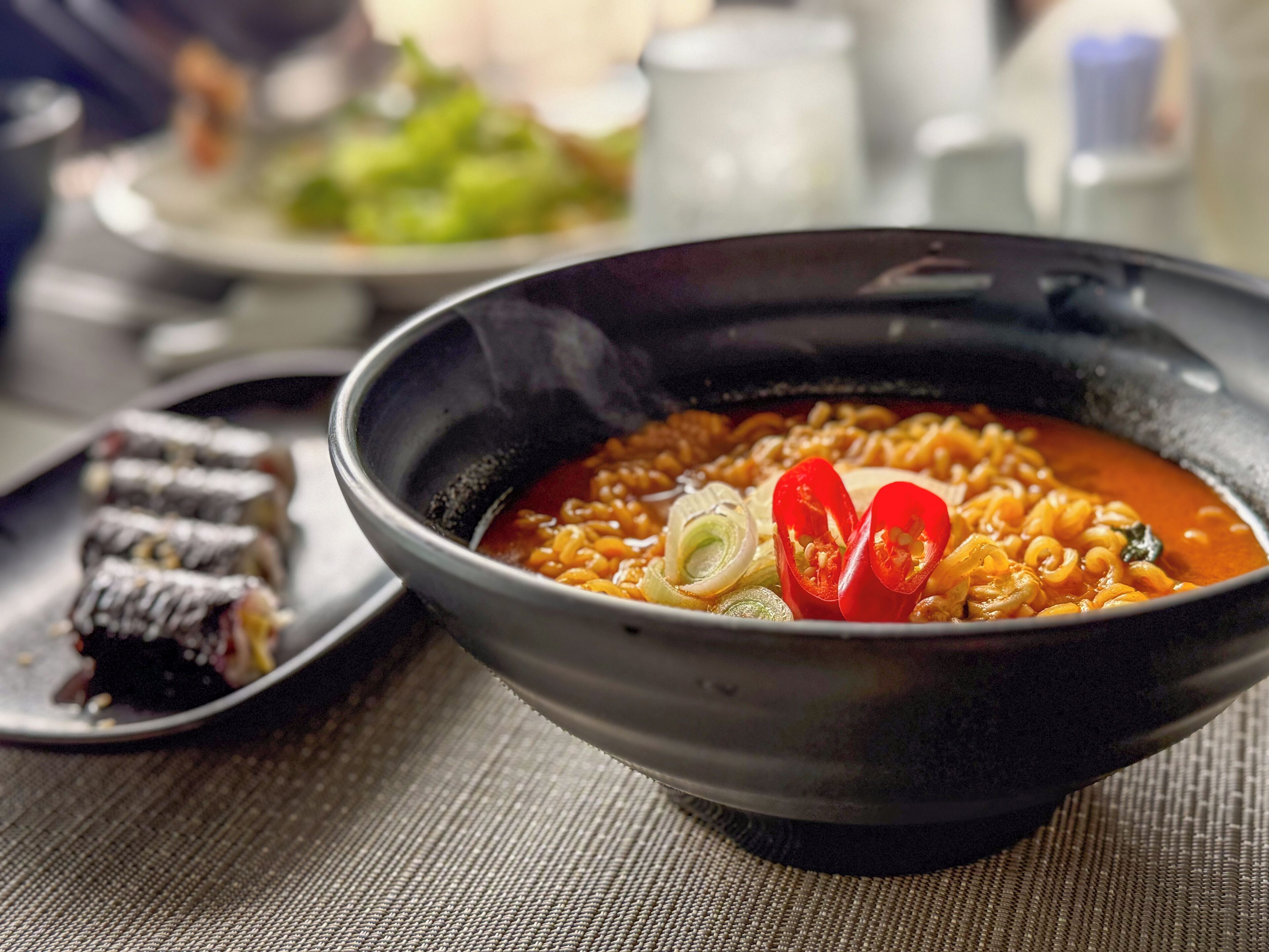 Un bol de ramen con ingredientes, un plato de vegetales encurtidos y un pequeño tazón de sushi sobre una mesa.