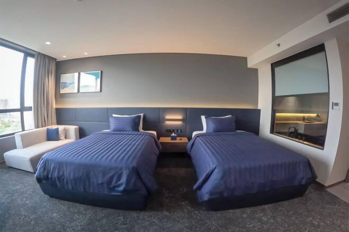 Ein Hotelzimmer mit zwei Betten, einem Schlafsofa, einem Nachttisch, einer Lampe und einem großen Fenster.