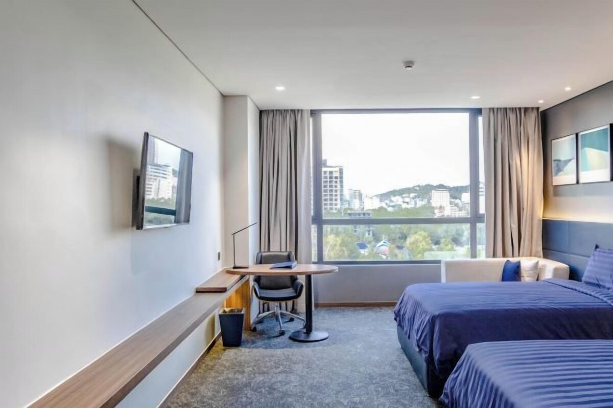 A hotel room with a bed, a desk, a chair, a TV, and a large window offering a city view.