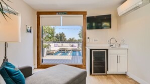 A compact room with a sofa, a small kitchenette, a TV mounted on the wall, and a view of an outdoor deck with a pool. - NEW BUILD Pet Friendly I Highlands I RiNo I Dwntwn (Denver)