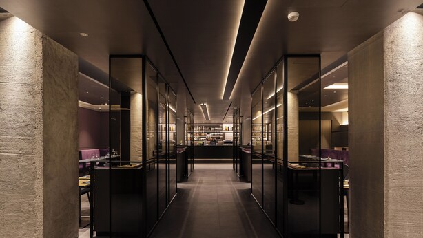 Un couloir d’hôtel moderne avec des cloisons en verre, un espace salon central et un bar visible au fond.
