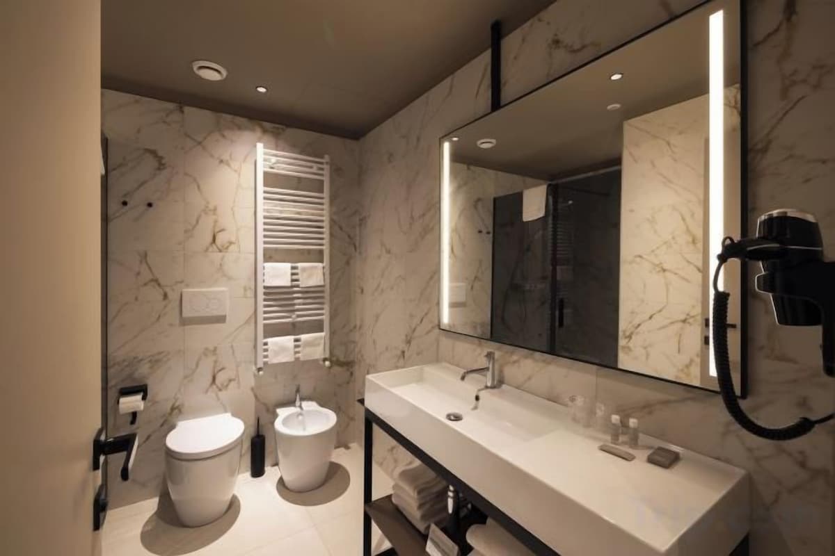 Une salle de bain moderne avec un mur en marbre, un grand miroir, des toilettes, un bidet et un meuble-lavabo avec un évier et un porte-serviettes.