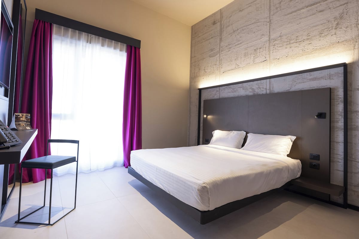 A modern hotel room with a large bed, a desk with a chair, and a view of the city through large windows with curtains.