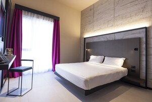 Camera d'hotel moderna con un letto grande, una scrivania con sedia e vista sulla città attraverso ampie finestre con tende.