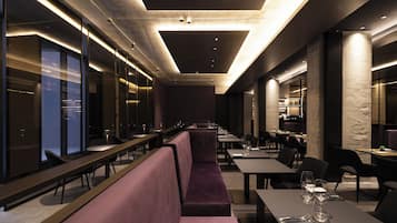 A modern restaurant with long communal tables, cushioned seating, and a high ceiling with recessed lighting.