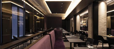 A modern restaurant with long communal tables, cushioned seating, and a high ceiling with recessed lighting.