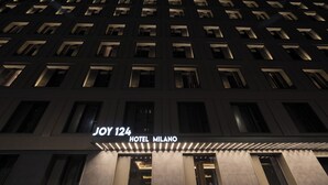 La façade d’un hôtel, avec des fenêtres illuminées et une enseigne indiquant « JOY 124 HOTEL MILANO ».