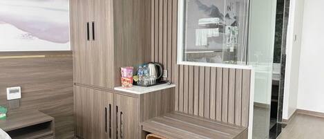 A modern hotel room with a wooden wardrobe, a small table with toiletries, a bench, and a glass shower enclosure.