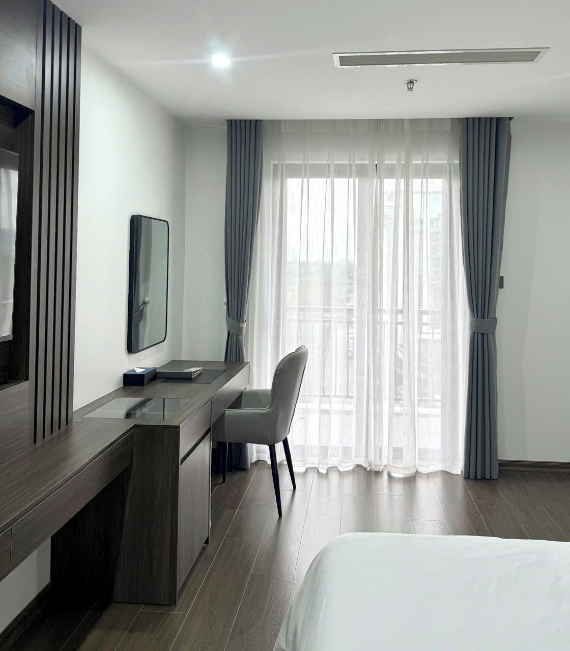 Una habitación de hotel moderna con cama, escritorio con silla, televisor de pantalla plana y ventanas amplias con cortinas translúcidas.