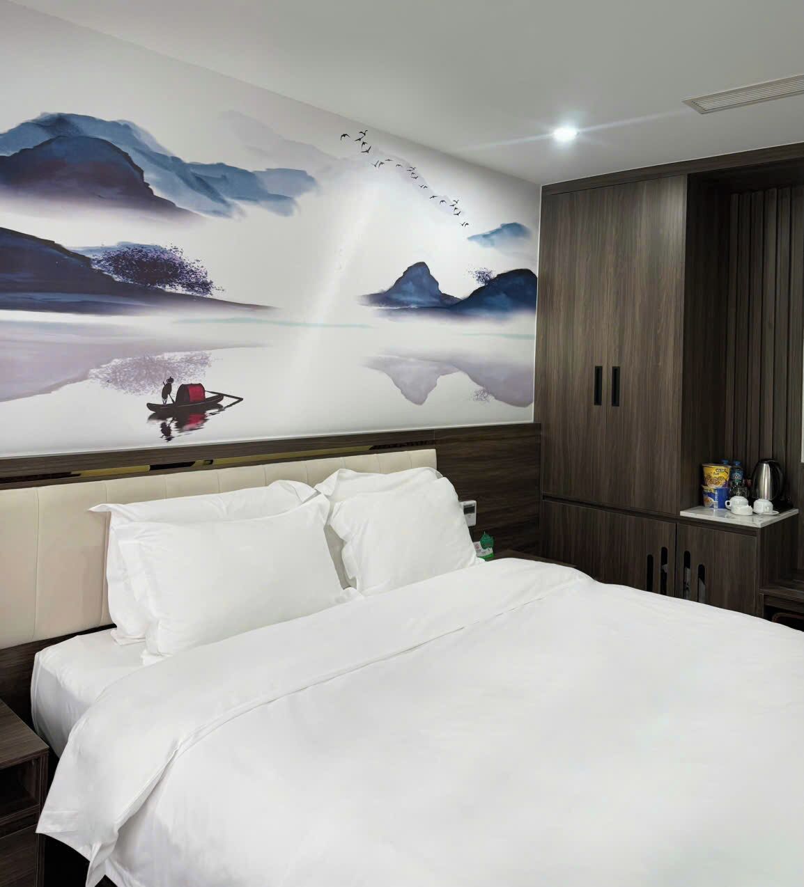 A hotel room with a large bed, a headboard with a mural, a wooden wardrobe, and a small table with a kettle and cups.