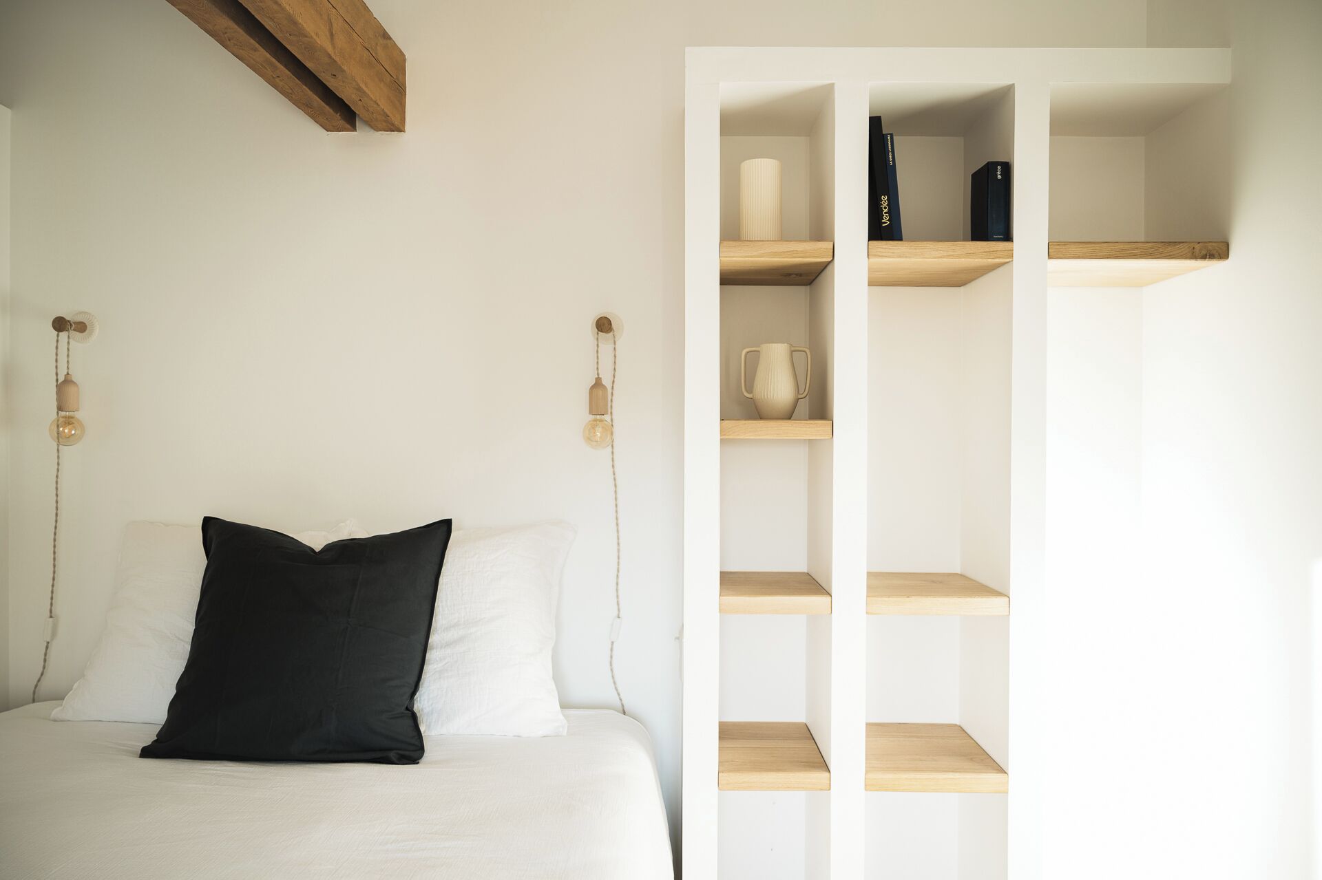 Een slaapkamer met een bed, een zwart kussen, een wit kussen, een houten dressoir met boeken en een vaas, en een wandlamp.