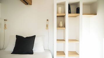 Een slaapkamer met een bed, een zwart kussen, een wit kussen, een houten dressoir met boeken en een vaas, en een wandlamp.