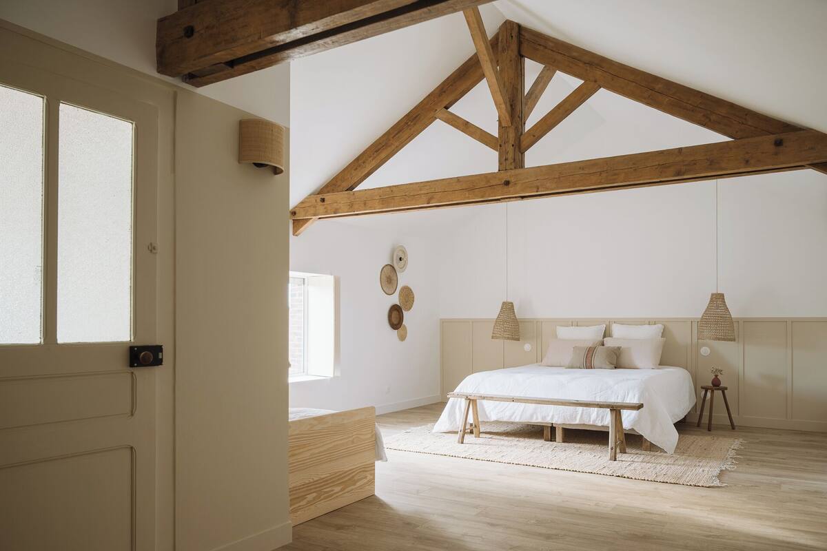 A bedroom with a wooden ceiling, a bed with white bedding, a bench, and a woven rug.