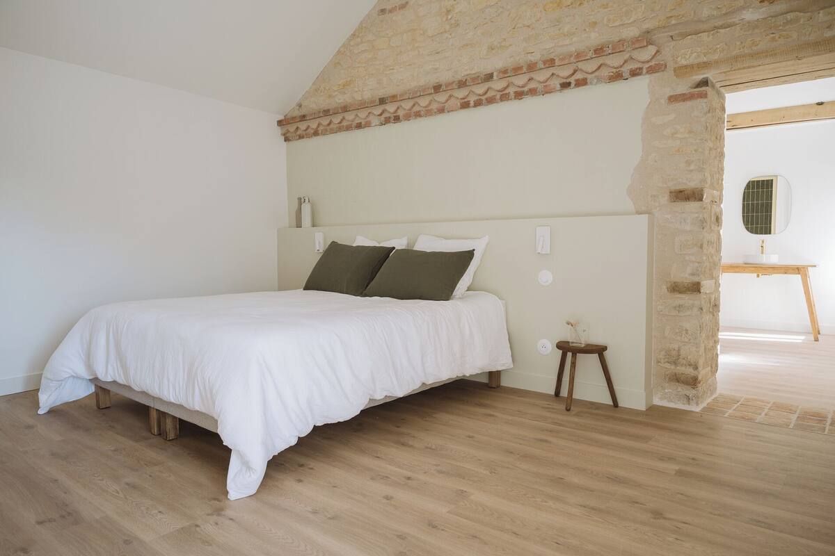 A bedroom with a wooden floor, a bed with white bedding, a stone wall, and a small wooden stool.