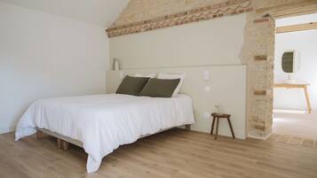 Un dormitorio con piso de madera, una cama con ropa blanca, una pared de piedra y un taburete de madera pequeño.