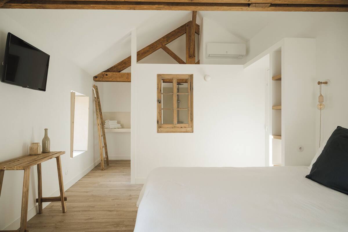 A bedroom with a wooden ceiling, a bed, a TV, a wooden ladder, a window, and a wall-mounted air conditioner.