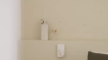A bedroom with a bed, a nightstand, a wall-mounted light, and a shelf with a bottle and dried flowers.