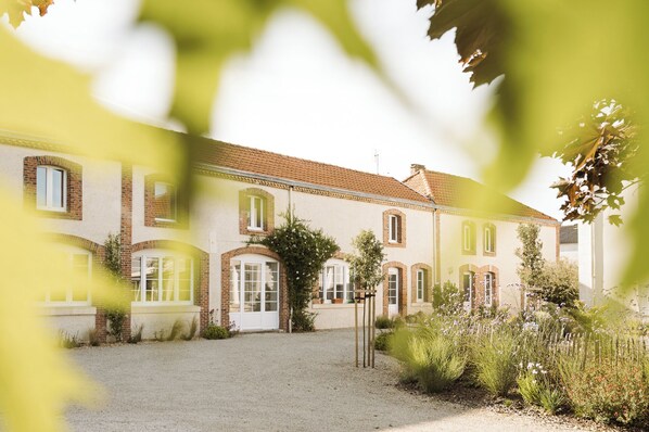 Un bâtiment à deux étages, avec une allée gravillonnée et un jardin agrémenté de diverses plantes et fleurs.