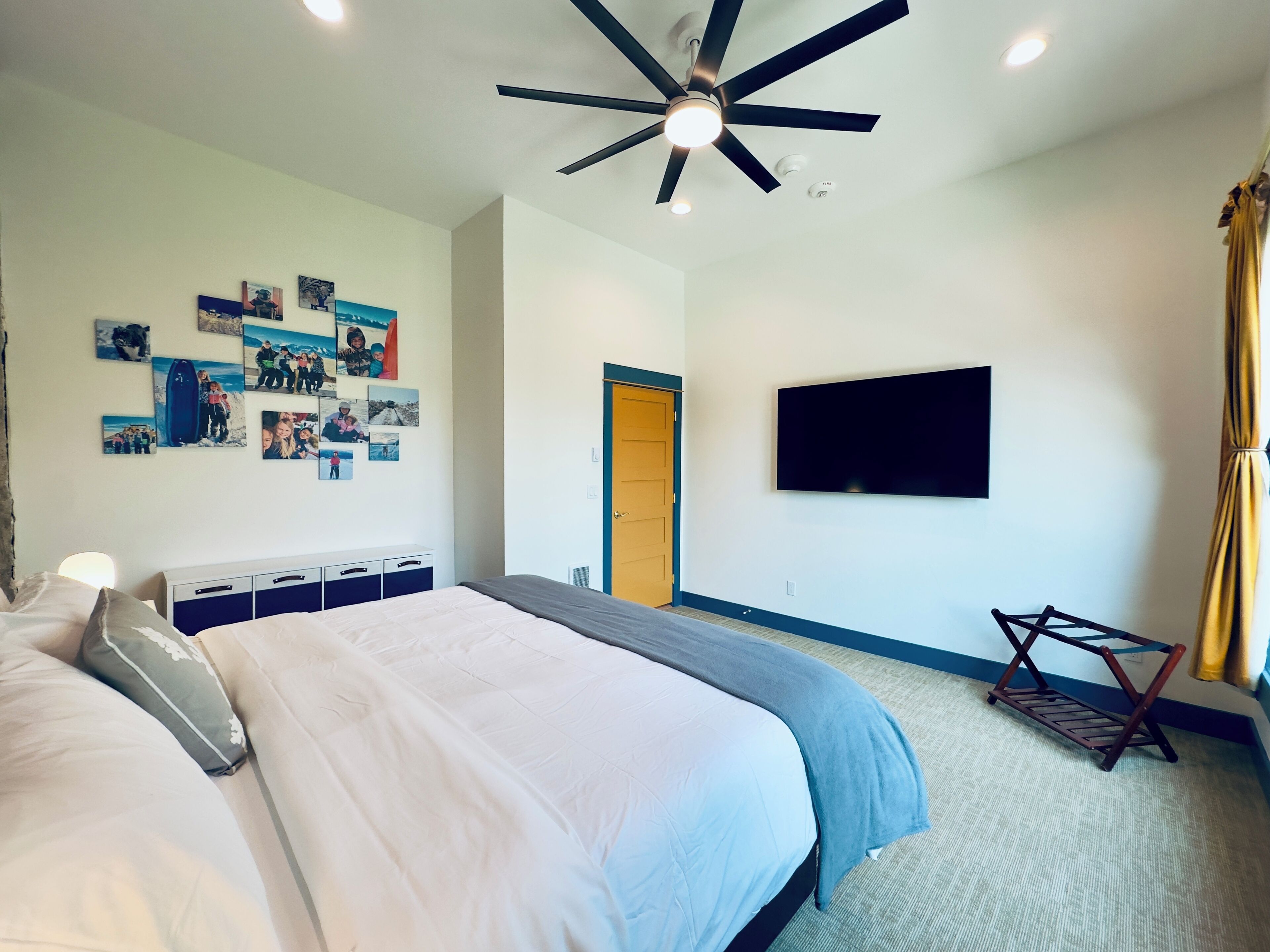 A bedroom with a bed, a ceiling fan, a mounted TV, and a door leading to another room.