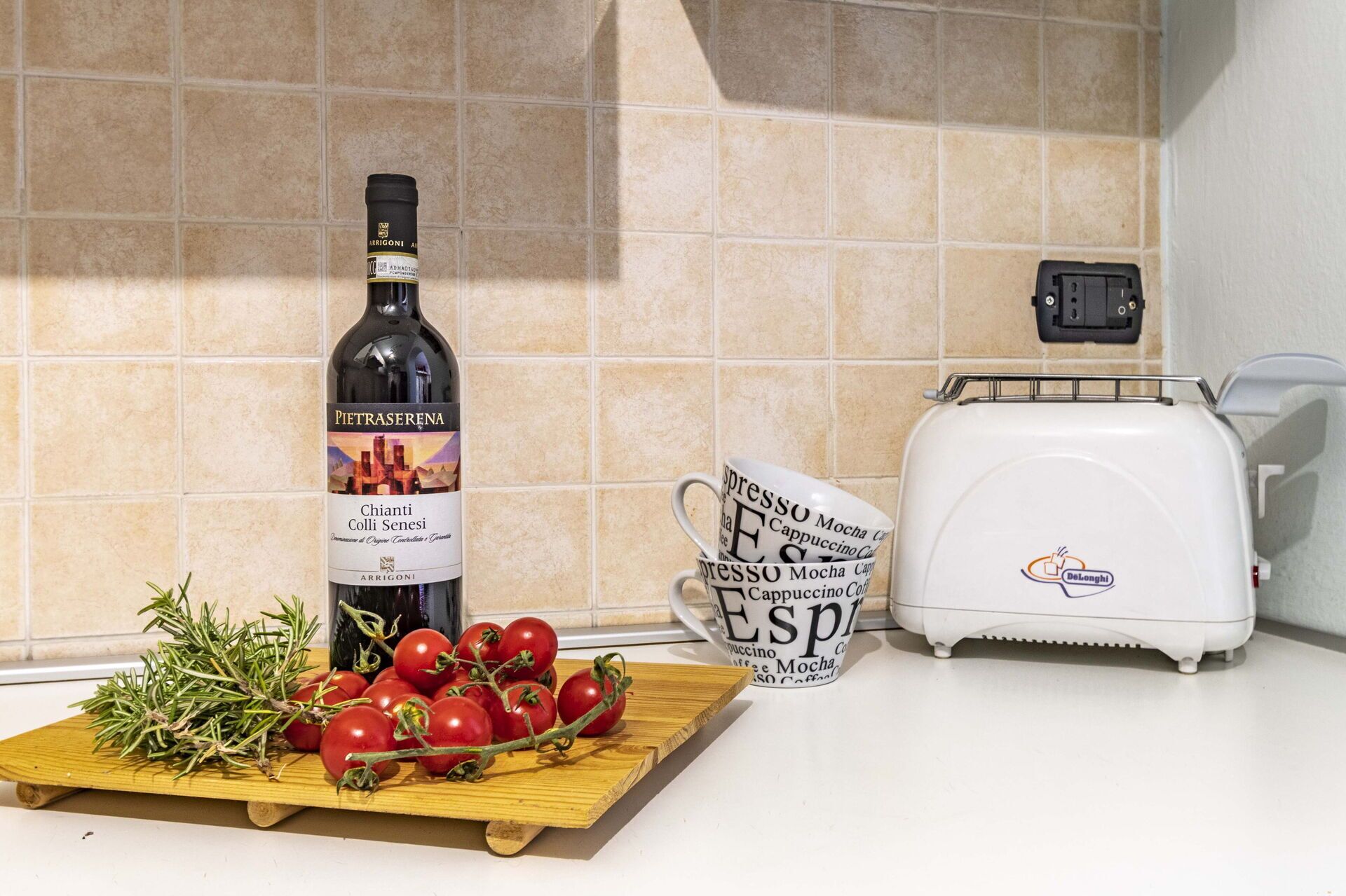 A kitchen counter with a bottle of wine, a cutting board with tomatoes and rosemary, and a coffee maker.
