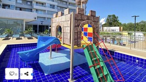Zona de juegos infantiles con tobogán y estructura de escalada, piscina con azulejos azules y un edificio con balcones al fondo.