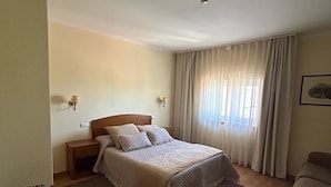 Une chambre d’hôtel avec un lit, une table de chevet, une chaise, une table ornée d’un vase de fleurs et une fenêtre avec des rideaux.