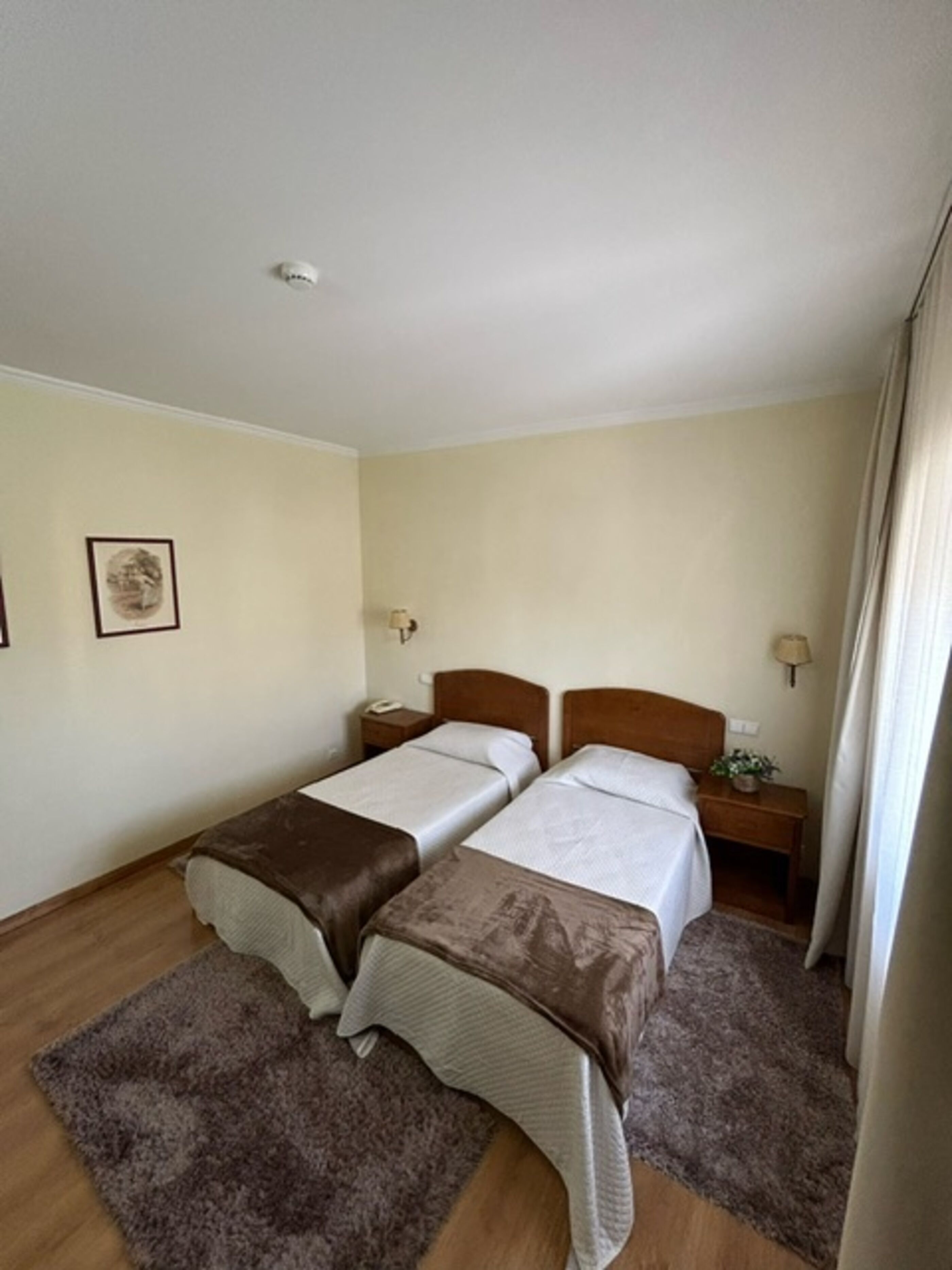 Una habitación con dos camas individuales, cabeceros de madera, mesitas de noche con lámparas, un cuadro enmarcado en la pared y una ventana con cortinas.