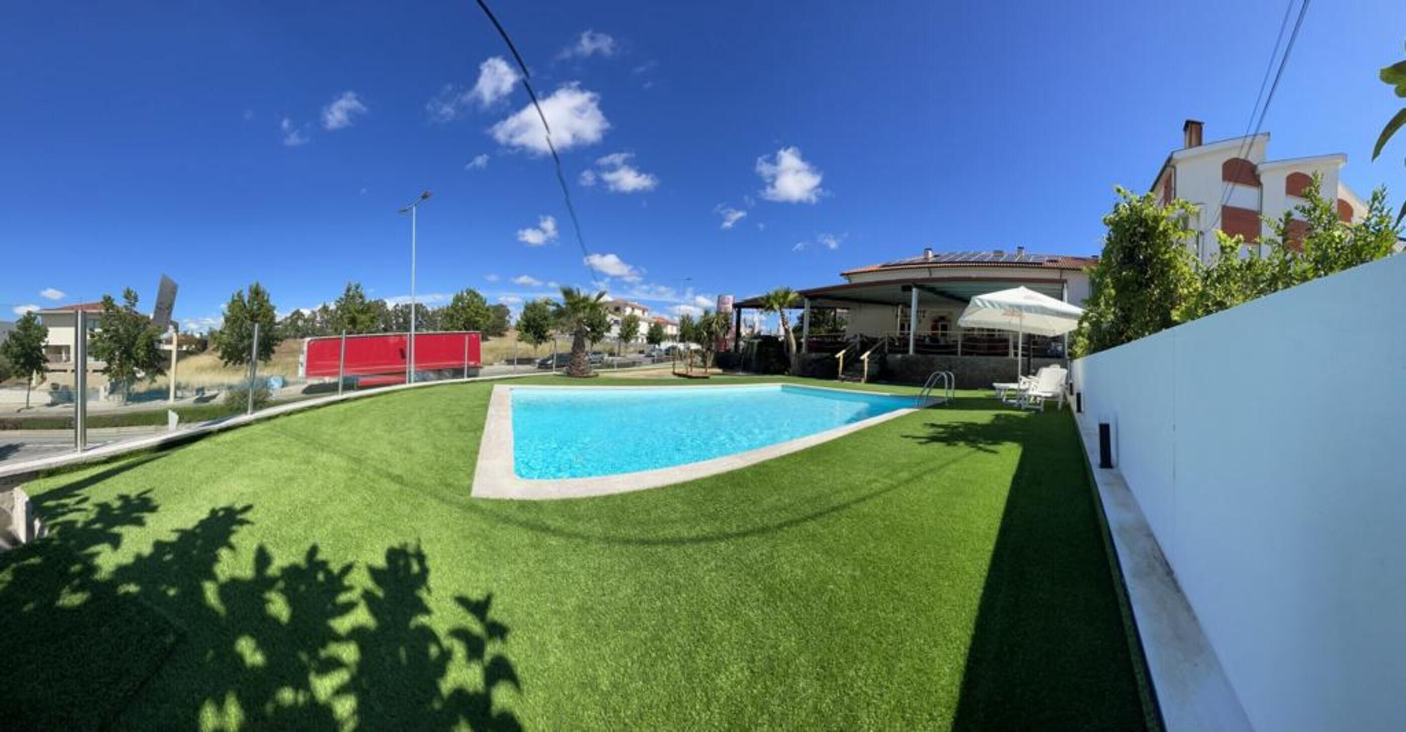 Una piscina rodeada de césped artificial, un porche cubierto y un edificio blanco con una estructura roja al fondo.