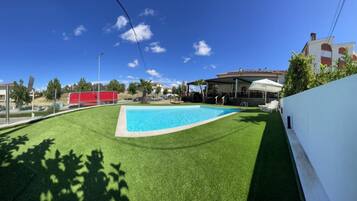 Una piscina rodeada de césped artificial, un porche cubierto y un edificio blanco con una estructura roja al fondo.