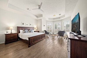 A bedroom with a large bed, a desk, a chair, a ceiling fan, and a cityscape TV.