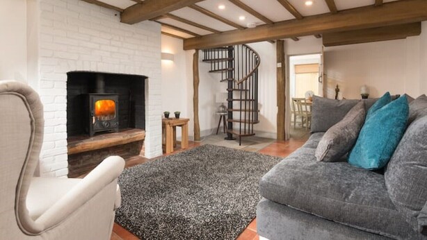 Una sala de estar acogedora con chimenea, un sofá gris con cojines azules y una escalera de caracol que lleva a otra habitación.
