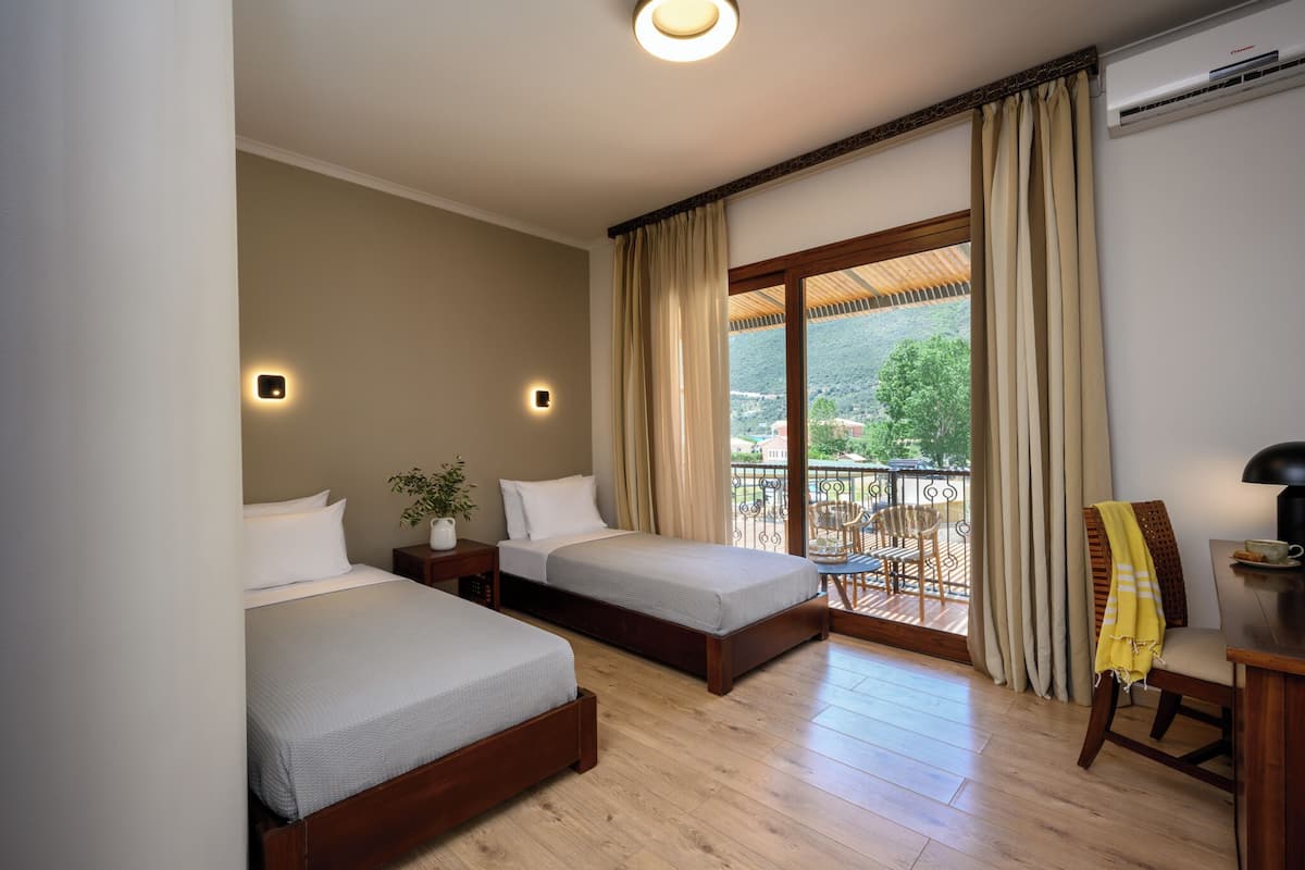 A hotel room with two single beds, a small table with a lamp, a chair, and a balcony with a view of greenery.