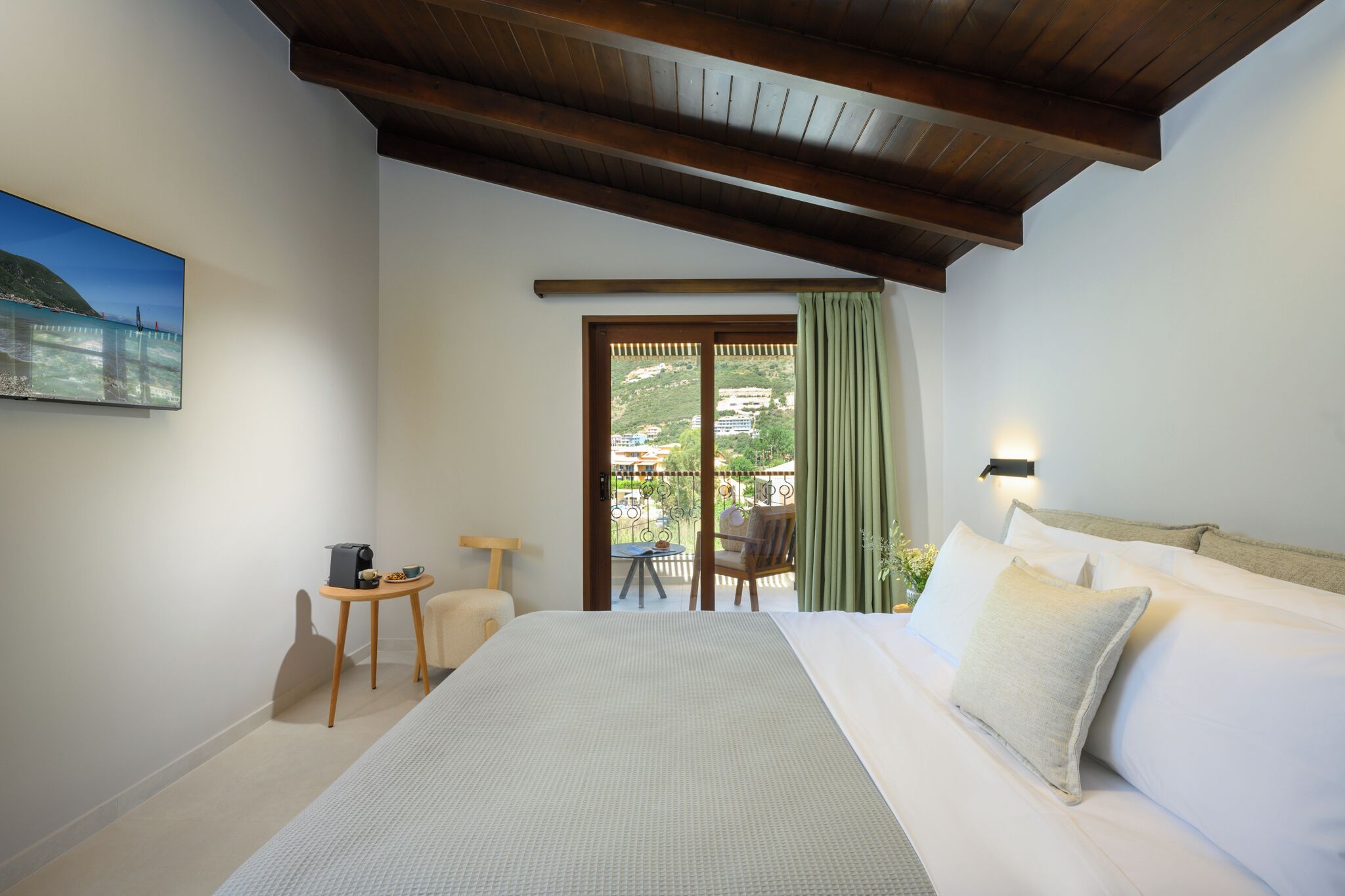 A hotel room with a bed, a small table with a chair, a painting on the wall, and a view of a balcony with a table and chairs.