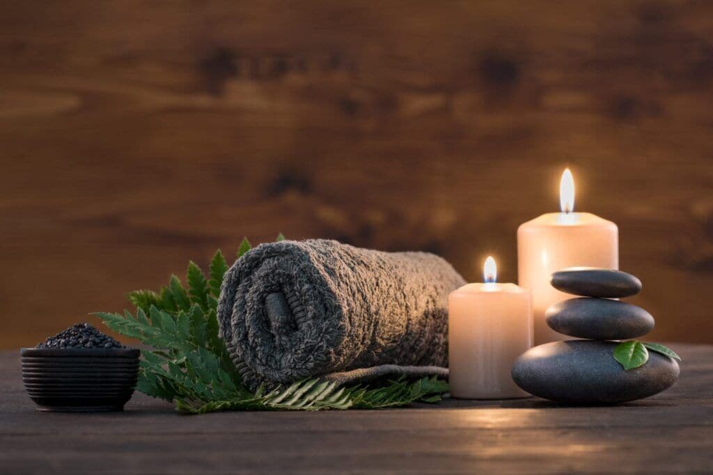 A wooden surface with a rolled towel, a lit candle, a stack of stones, and a bowl of black pebbles.
