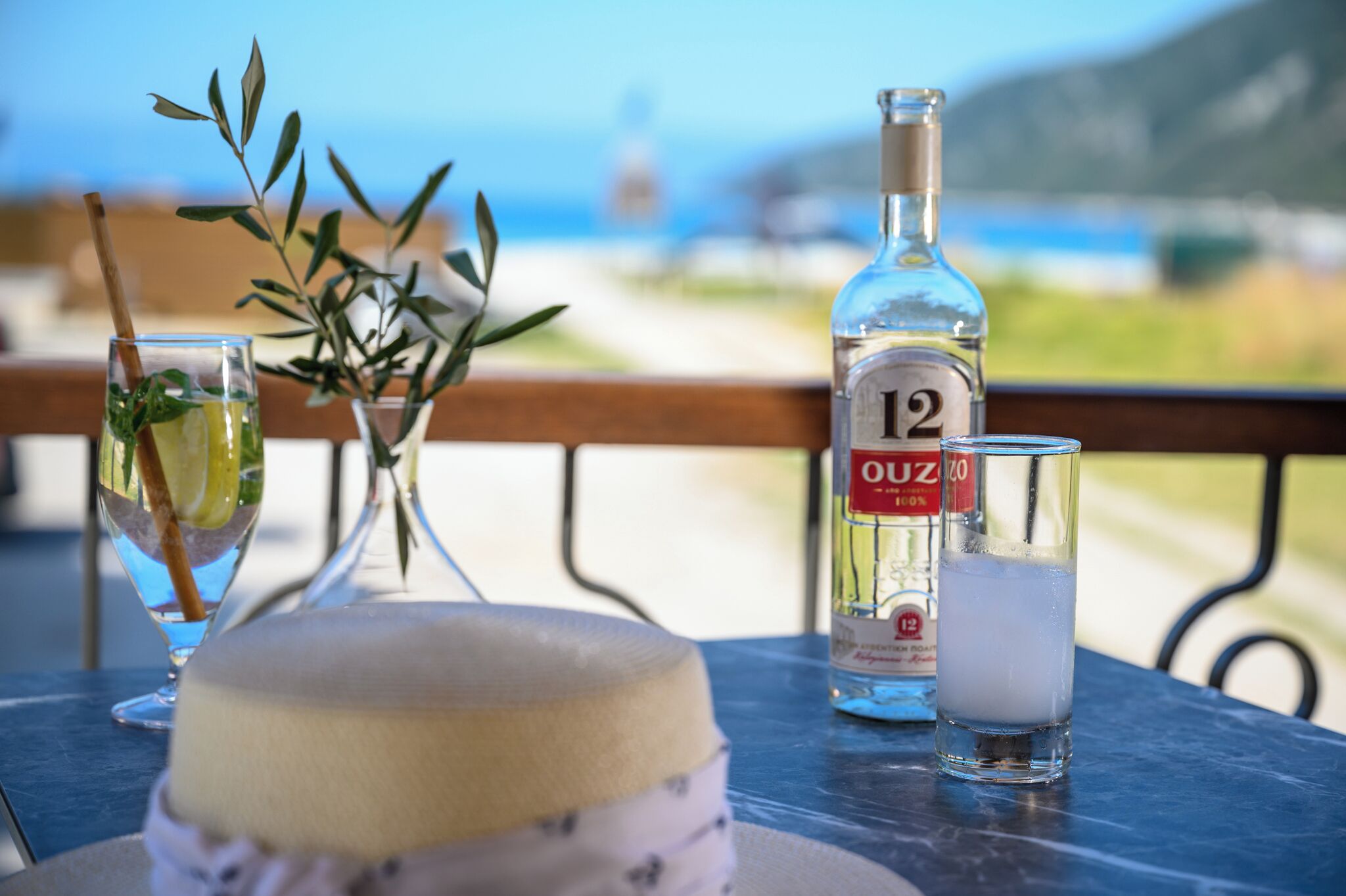 A table with a bottle of liquor, a glass with ice, and a dessert on a balcony overlooking a scenic view.