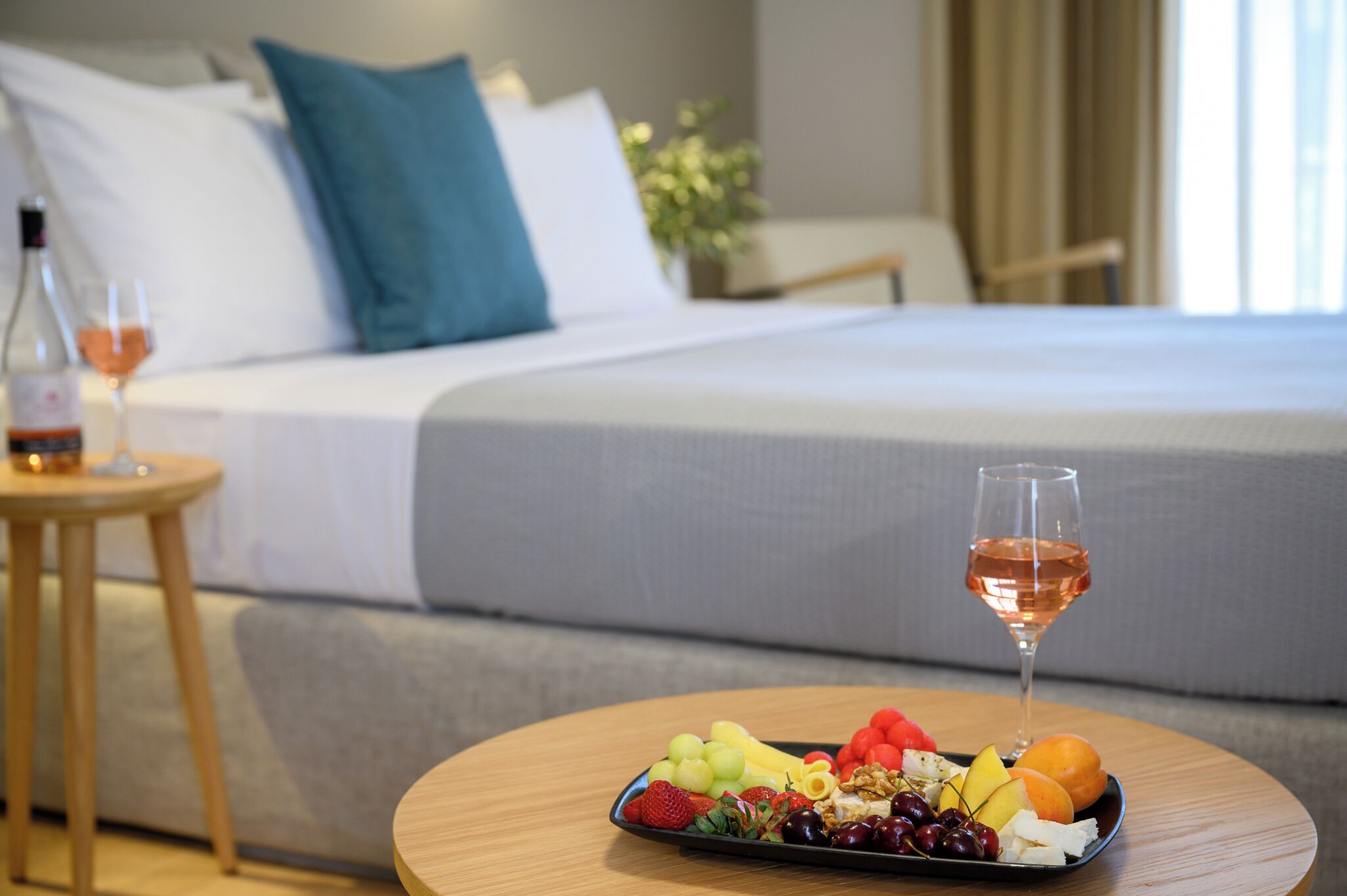 A neatly made bed with white and teal pillows, a bedside table with a bottle and glass, and a fruit platter on a round table in a hotel room.