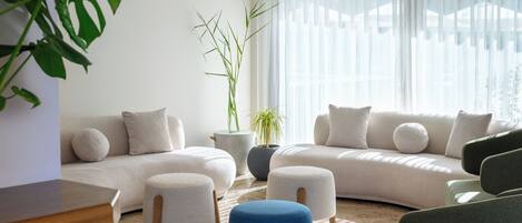 Un salon moderne avec un canapé beige, un pouf bleu et un fauteuil gris. On y trouve une grande plante en pot et une petite table avec un plateau en bois.