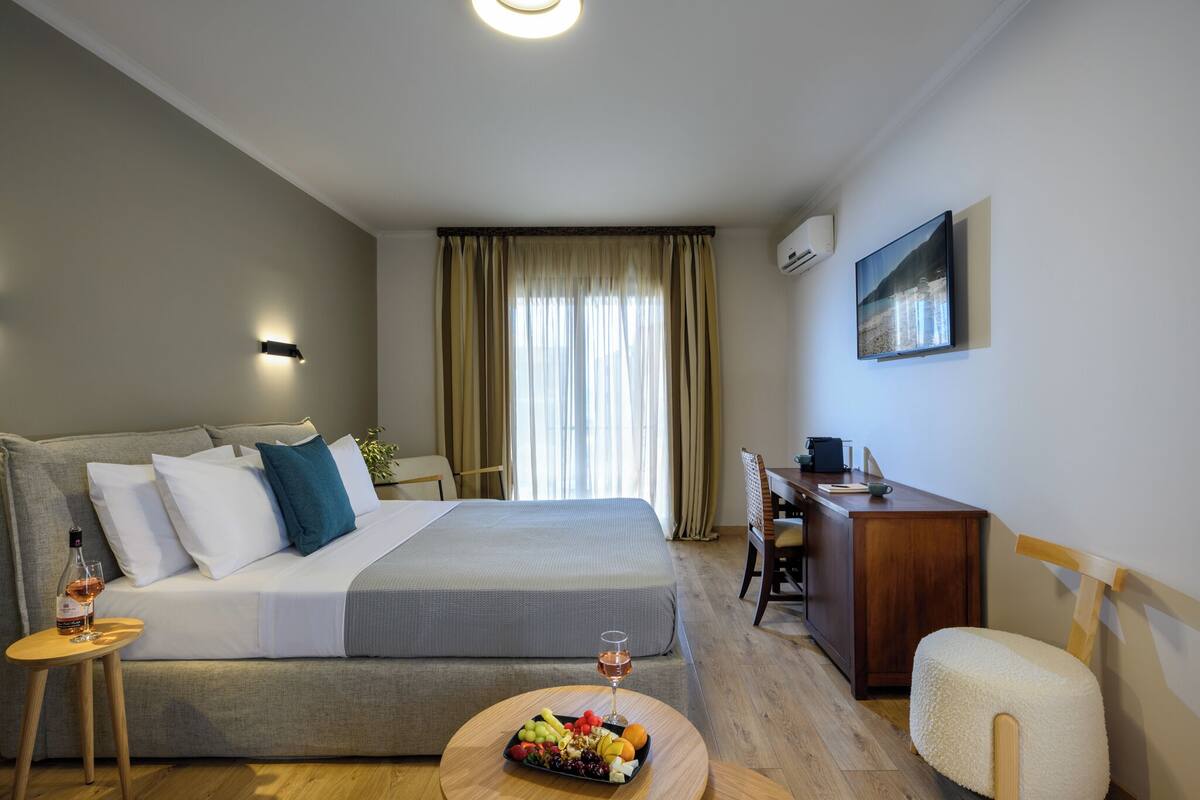 A hotel room with a bed, a desk, a chair, a small table with fruit and a glass of wine, and a painting on the wall.
