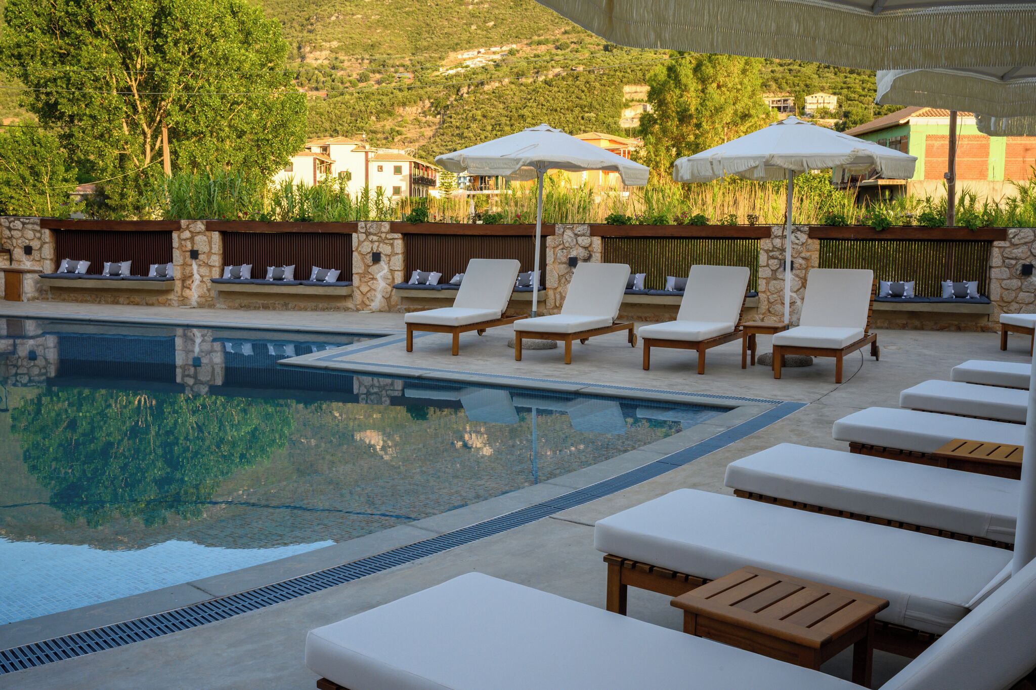 A poolside area with lounge chairs and umbrellas, stone walls, and a view of buildings in the background.