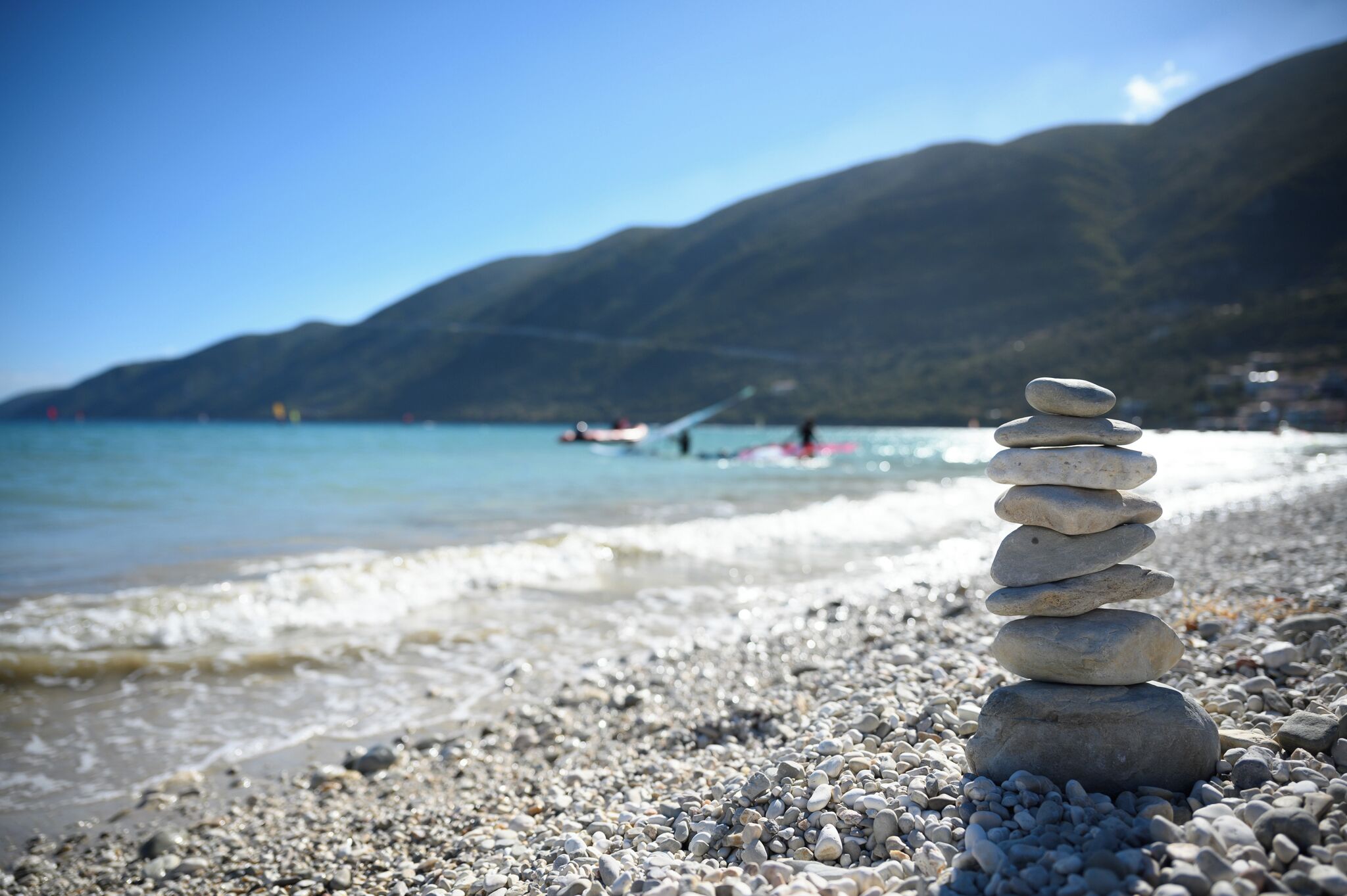 A stack of stones on a pebbly beach with a calm sea and mountains in the background.