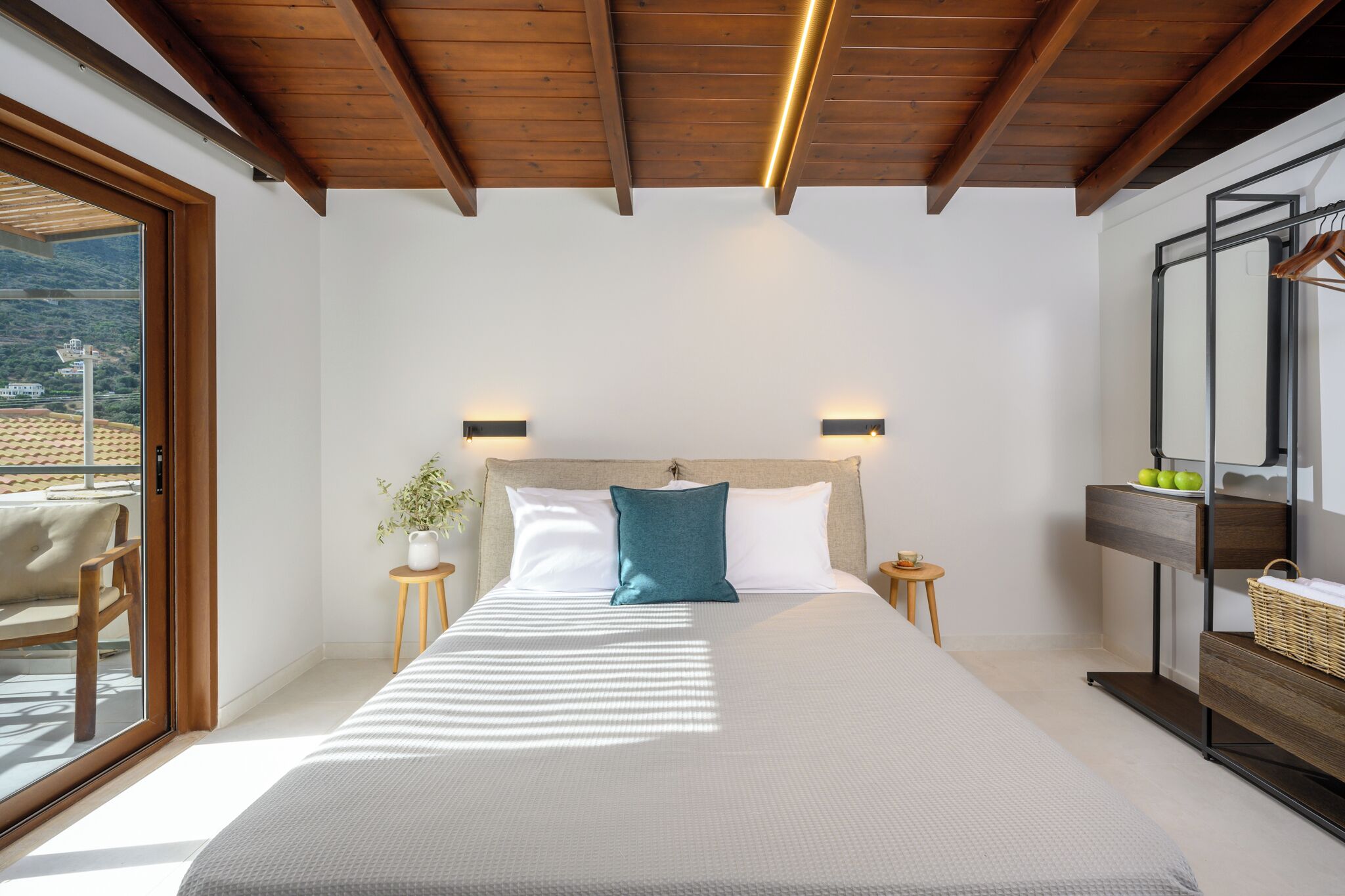 Une chambre avec un grand lit, des poutres apparentes en bois au plafond, un balcon avec une chaise et une vue sur un immeuble.