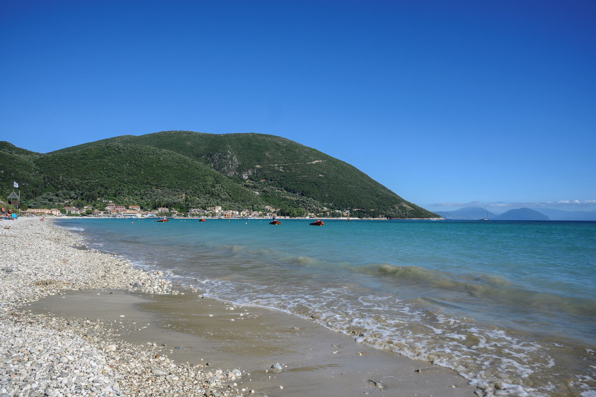 A pebble beach with clear turquoise water, a green hill in the background, and a sailboat in the distance.