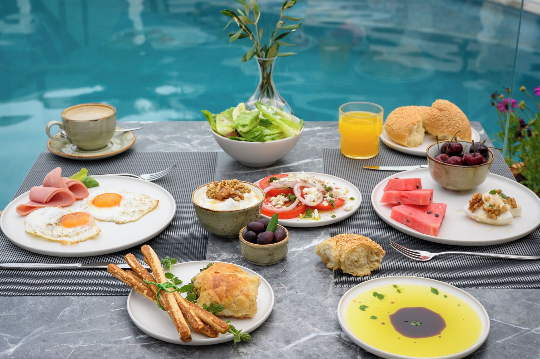 A breakfast setup by a pool with plates of eggs, toast, and fruit, along with a cup of coffee and a glass of orange juice.
