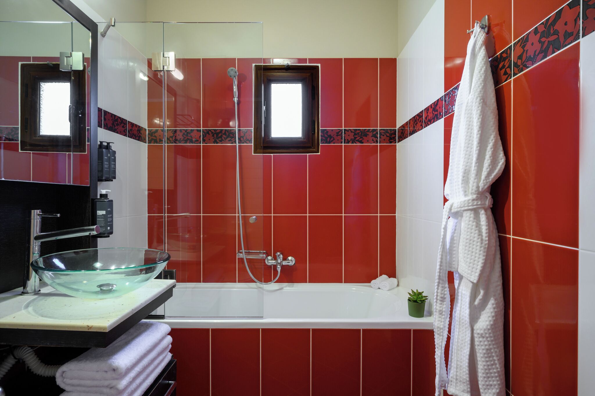 A bathroom with a glass bowl sink, a white bathtub, and a red tiled wall.