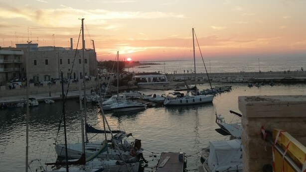 Un puerto deportivo con barcos amarrados, un atardecer al fondo y edificios a lo largo de la costa.