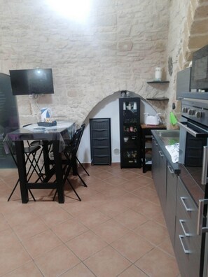 A kitchen with a stone wall, a dining table with chairs, a TV mounted above the table, and a built-in oven. - Gorgeous stone built apartment in the heart of Giovinazzo's historic center (Giovinazzo)