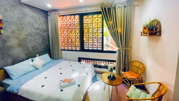 A hotel room with a bed, a small round table, two wicker chairs, a window with curtains, and a shelf with a plant. - Mango Home & Apartment (Da Nang)